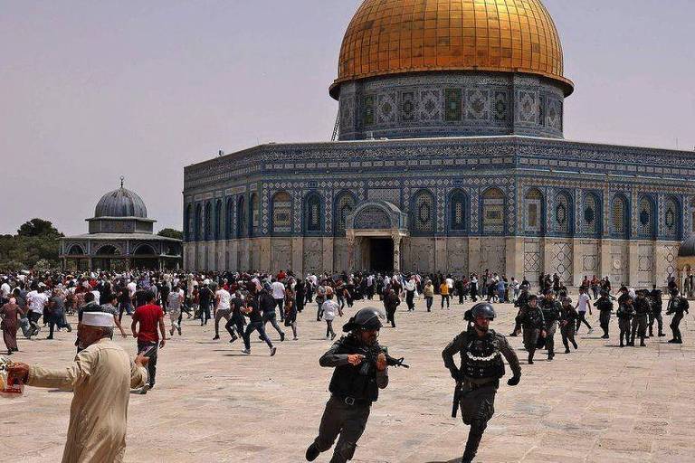 A imagem mostra uma cena de tumulto em um local sagrado em Jerusalém, com uma grande multidão de pessoas correndo. No centro, destaca-se uma grande cúpula dourada, que é parte de uma estrutura arquitetônica significativa. Policiais em uniformes estão presentes, alguns correndo, enquanto outras pessoas parecem estar em movimento rápido, sugerindo uma situação de tensão. O céu está claro e a cena é diurna.