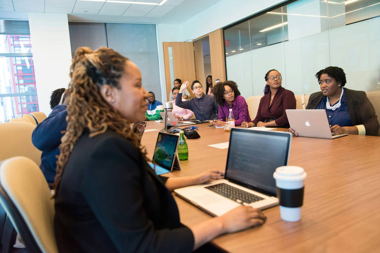 A imagem mostra um grupo de pessoas em uma sala de conferência. À frente, uma mulher com cabelo cacheado e preso está usando um laptop. Ao fundo, outras mulheres estão sentadas ao redor de uma mesa, algumas com laptops e outras com tablets. A sala é bem iluminada, com janelas grandes e paredes de vidro. Há também garrafas de água e copos sobre a mesa.