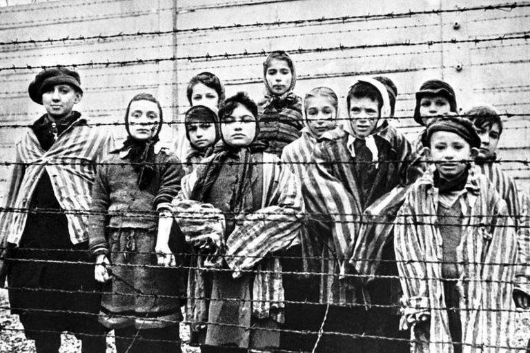A imagem mostra um grupo de crianças em um campo de concentração, vestindo roupas listradas. Elas estão posicionadas em frente a uma cerca de arame farpado, com uma parede ao fundo. As crianças têm expressões sérias e estão agrupadas, algumas com lenços na cabeça. A fotografia é em preto e branco, transmitindo um sentimento de tristeza e desolação.