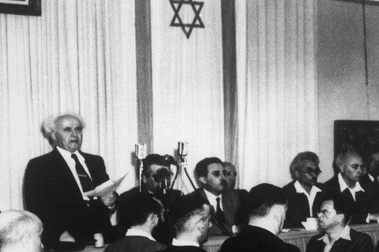 A imagem mostra uma reunião em um ambiente formal, com várias pessoas sentadas em uma mesa. Um homem está de pé, segurando papéis e falando, enquanto os outros o escutam atentamente. Ao fundo, há uma bandeira com a Estrela de Davi e cortinas brancas. A cena é em preto e branco, sugerindo que foi tirada em uma época passada.