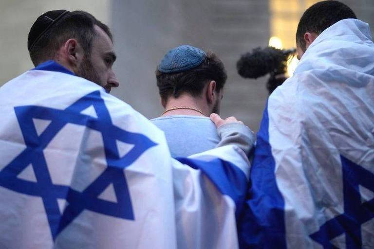 Três homens estão de costas, envoltos em bandeiras de Israel. Um deles usa um quipá. Eles parecem estar em um ambiente de manifestação ou evento, com um microfone visível em uma das mãos. A cena transmite um sentimento de união e identidade cultural.