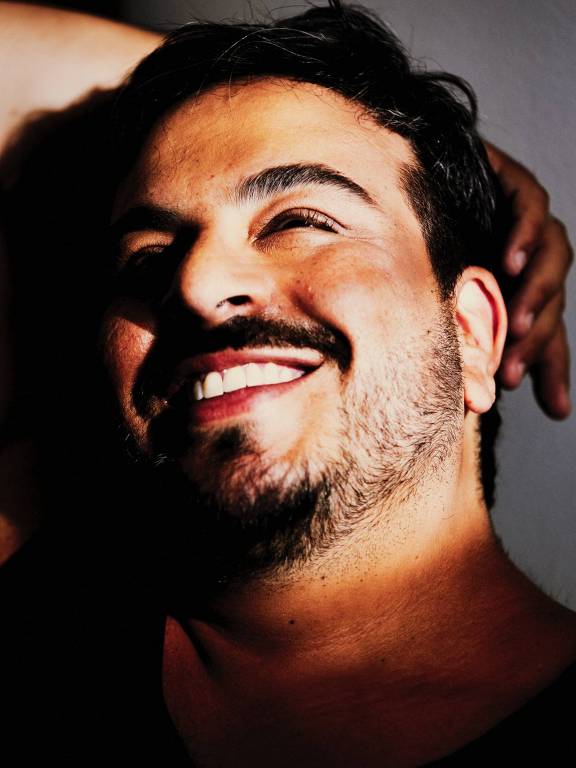 Um homem com cabelo escuro e barba está sorrindo. Ele está posicionado de lado, com uma mão atrás da cabeça e a outra ao lado do rosto. A luz incide sobre seu rosto, destacando seu sorriso e criando sombras. O fundo é neutro e claro, o que faz o homem se destacar na imagem.
