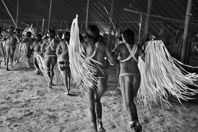 A imagem em preto e branco mostra um grupo de pessoas dançando em um ambiente rústico. As figuras estão de costas, vestindo trajes tradicionais e adornos feitos de palha ou material similar, com longas franjas que se destacam. O chão é de terra e o espaço parece ser uma estrutura coberta, possivelmente uma maloca ou espaço de celebração indígena.