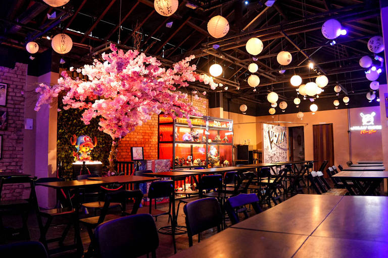 A imagem mostra o interior de um restaurante com mesas e cadeiras dispostas de forma organizada. No centro, há uma árvore de flores rosa, que se destaca na decoração. O ambiente é iluminado por lanternas penduradas no teto, criando uma atmosfera aconchegante. Ao fundo, há prateleiras com objetos decorativos e uma parede com arte. O espaço parece ser moderno e convidativo.