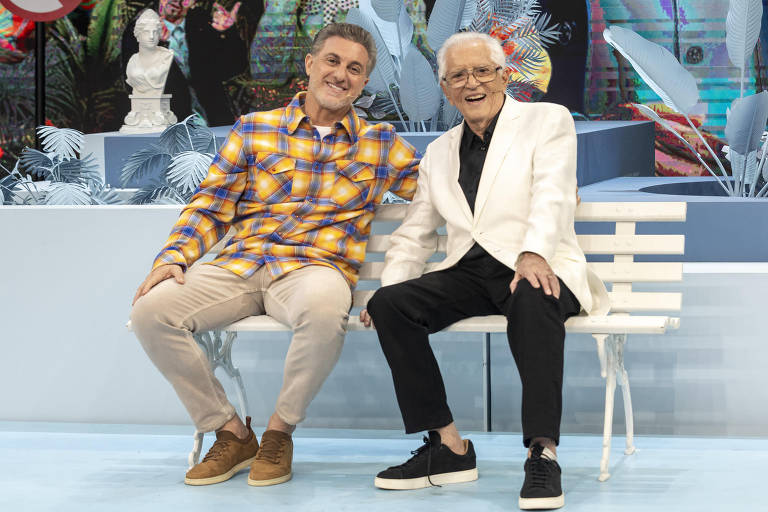 Dois homens estão sentados em um banco branco em um cenário de programa de TV. O homem à esquerda usa uma camisa xadrez colorida e calças claras, enquanto o homem à direita está vestido com um terno branco e calças escuras. Ambos estão sorrindo e parecem estar se divertindo. Ao fundo, há elementos decorativos como plantas e uma tela com imagens coloridas.
