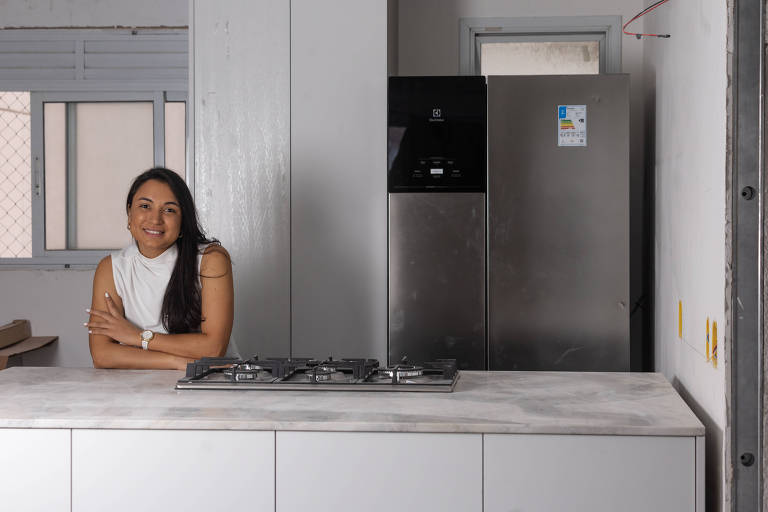 Een vrouw zit aan een modern aanrecht, met een gasfornuis ervoor. Op de achtergrond is een zilveren koelkast en een raam met tralies te zien. De vrouw heeft lang, steil haar en draagt een witte blouse. De omgeving is goed verlicht en heeft lichte muren.