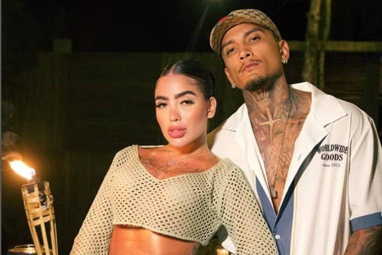 A imagem mostra um casal posando em um ambiente externo à noite, com iluminação suave. A mulher está vestindo um conjunto de crochê que destaca seu corpo tonificado e segura uma tocha. O homem está ao seu lado, usando uma camisa de estilo casual e com tatuagens visíveis. O fundo apresenta elementos de decoração ao ar livre, como luzes e madeira.
