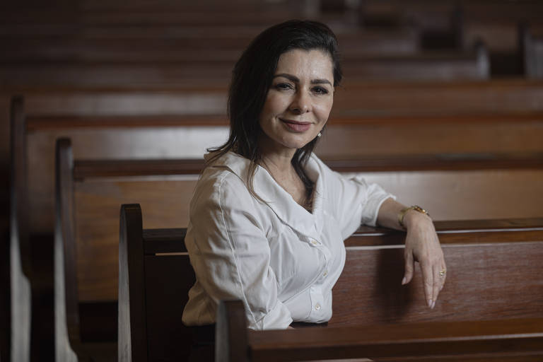 Uma mulher está sentada em um banco de madeira em um ambiente que parece ser uma igreja. Ela está vestindo uma camisa branca e tem cabelo escuro e liso. A mulher sorri enquanto apoia o braço em um dos bancos. Ao fundo, há vários bancos de madeira dispostos em fileiras, criando uma atmosfera tranquila e serena.