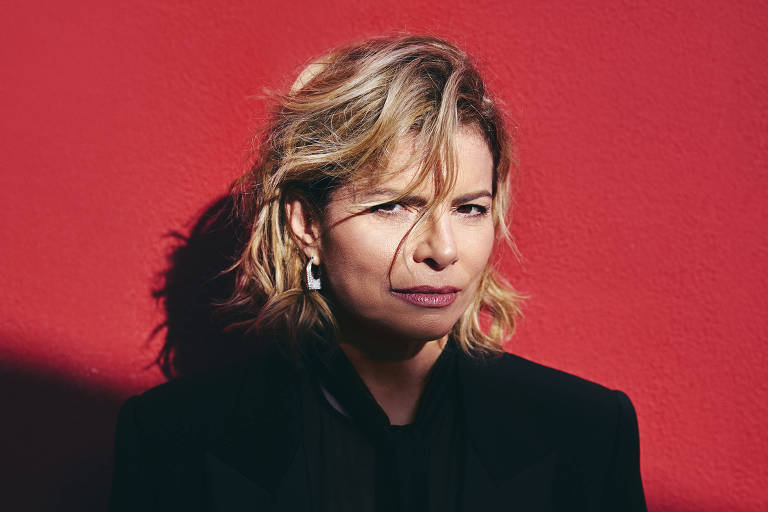 A imagem mostra uma mulher com cabelo curto e ondulado, usando um blazer preto. Ela está em pé em frente a uma parede vermelha, com uma sombra projetada sobre ela. A expressão dela é séria e ela olha diretamente para a câmera.