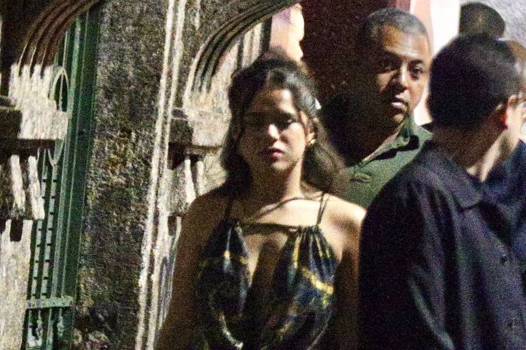 A imagem mostra uma mulher caminhando em uma rua à noite. Ela está vestindo um vestido curto com um padrão colorido e sapatos de salto alto. Ao fundo, há outras pessoas, e a iluminação é baixa, sugerindo um ambiente noturno. A mulher parece confiante enquanto caminha.