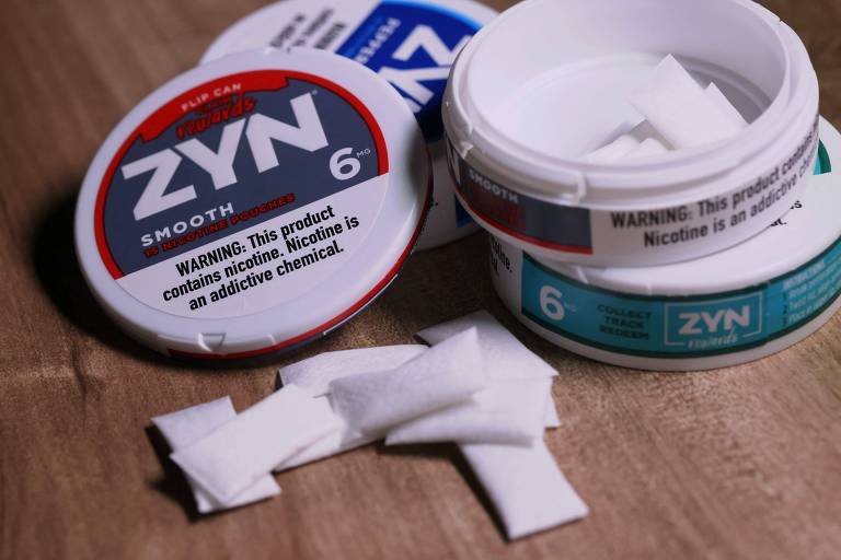 A imagem mostra duas latas de produtos ZYN, uma delas aberta, com várias pequenas porções brancas visíveis. As latas são redondas e possuem rótulos que indicam o nome 'ZYN' e o número '6'. Uma das latas contém um aviso sobre a presença de nicotina, que é uma substância química viciante. O fundo da imagem é uma superfície de madeira.