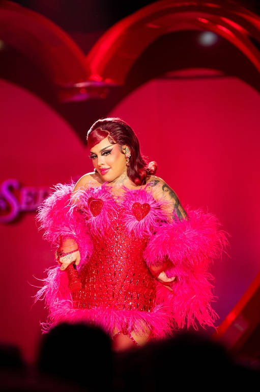 Uma pessoa está se apresentando em um desfile de moda, vestindo um traje vermelho com detalhes em rosa e penas. O fundo é decorado com corações e luzes, criando uma atmosfera vibrante e festiva. A pessoa tem cabelo rosa e está posando de forma confiante, com um sorriso no rosto.