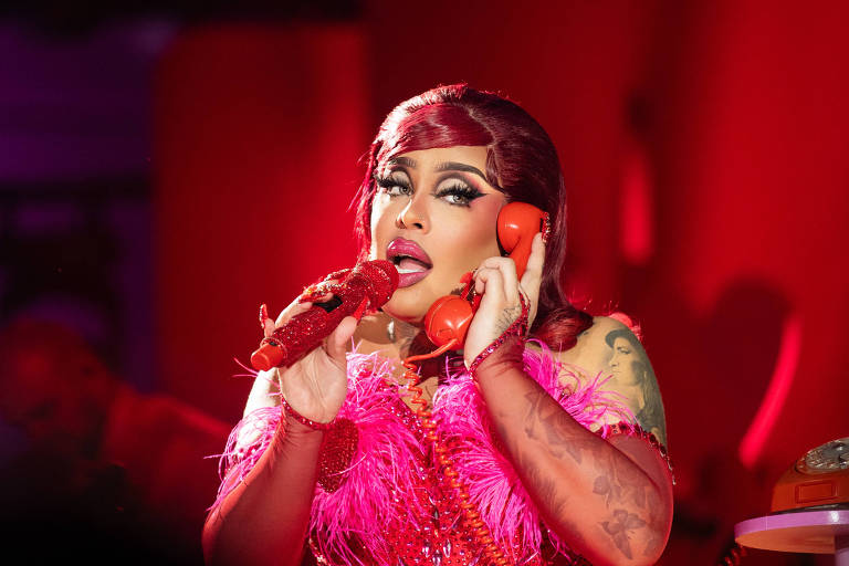 Uma artista drag está se apresentando em um palco iluminado com luzes vermelhas. Ela usa um vestido brilhante e rosa, decorado com penas. A artista segura um telefone vermelho em uma mão e parece estar cantando ou se comunicando. O fundo é desfocado, mas sugere um ambiente de show com público.