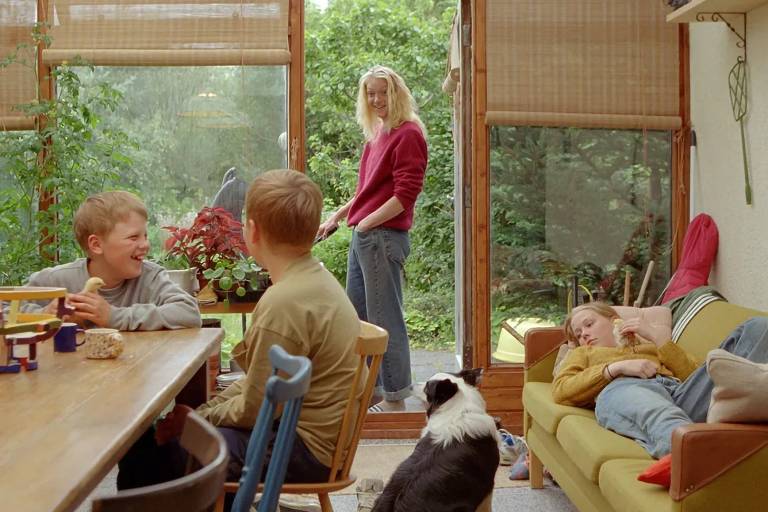 A imagem mostra um ambiente interno com uma mesa de madeira onde duas crianças estão sentadas, sorrindo e conversando. Uma terceira criança está deitada em um sofá, enquanto uma mulher com cabelo longo e loiro está em pé perto de uma porta de vidro, olhando para o exterior. Um cachorro preto e branco está no chão, próximo à mesa. O ambiente é iluminado e possui plantas ao fundo, visíveis através da porta de vidro.