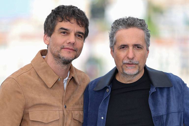 A imagem mostra dois homens posando juntos. O homem à esquerda tem cabelo cacheado e usa uma camisa de couro clara. O homem à direita tem cabelo grisalho e usa uma jaqueta escura. O fundo é desfocado, sugerindo um ambiente ao ar livre.