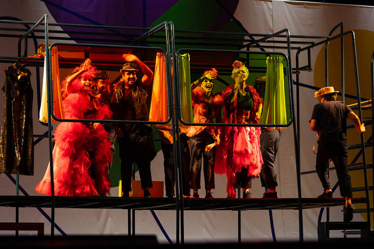 A imagem mostra um grupo de pessoas em um cenário teatral. Elas estão em uma estrutura metálica, com roupas coloridas e extravagantes, incluindo um vestido volumoso rosa. Algumas pessoas estão acenando e sorrindo, enquanto outras parecem estar se preparando para se apresentar. Ao fundo, há um pano de fundo colorido com formas geométricas. A iluminação é vibrante, destacando as cores das roupas e do cenário.