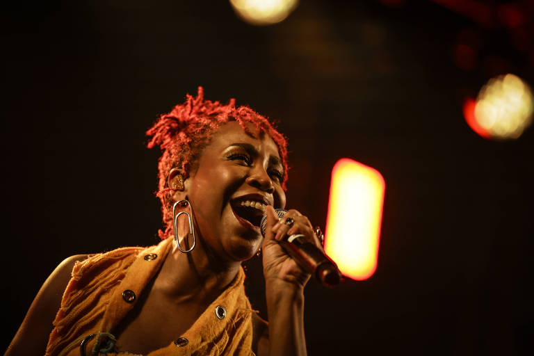 Uma mulher com cabelo curto e encaracolado, tingido de vermelho, está cantando em um microfone. Ela usa um colete amarelo e brincos grandes. O fundo é desfocado, com luzes coloridas, principalmente em tons de vermelho e amarelo, criando uma atmosfera de show ao vivo.