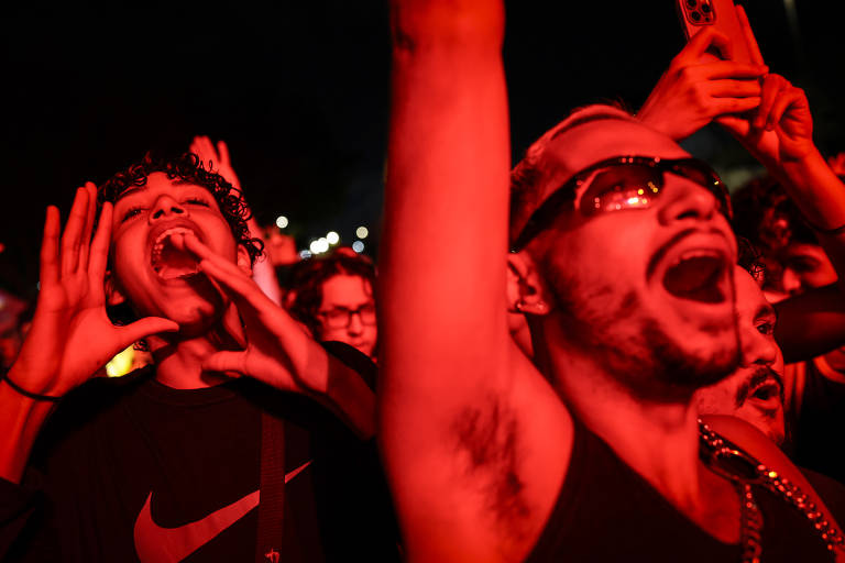 A imagem mostra um grupo de pessoas em um show, com foco em dois homens na frente. Um deles, à esquerda, tem cabelo cacheado e está gritando, enquanto o outro, à direita, usa óculos escuros e também está gritando. A iluminação é predominantemente vermelha, criando uma atmosfera vibrante e energética. Ao fundo, outras pessoas podem ser vistas, algumas segurando celulares.