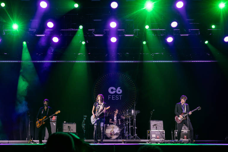 A imagem mostra uma banda se apresentando em um palco iluminado com luzes verdes e roxas. Os membros da banda incluem um guitarrista à esquerda, um vocalista e guitarrista ao centro, um baterista ao fundo e um guitarrista à direita. O fundo exibe o logo do evento 'C6 FEST'.