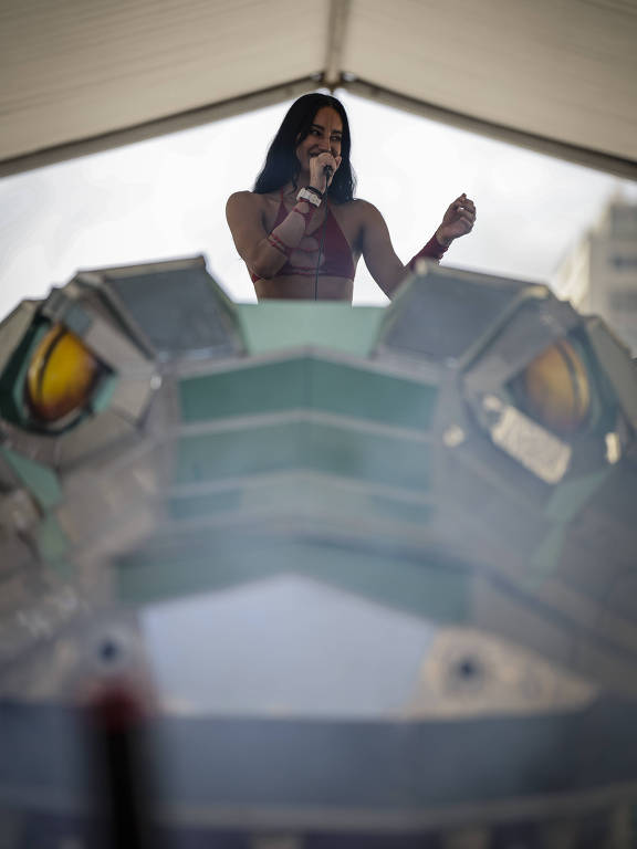 Uma artista está se apresentando em um palco elevado, que tem a forma de uma cabeça de dragão. Ela está usando um top vermelho e segurando um microfone, enquanto canta. O fundo é uma estrutura coberta, e há uma leve névoa ao redor, criando um efeito atmosférico.