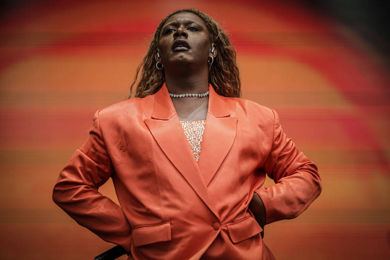 Uma artista se apresenta em um palco, vestindo um terno laranja. Ela está com as mãos na cintura e olhando para cima, transmitindo uma expressão de confiança. O fundo é desfocado, com cores quentes que sugerem um ambiente de show.