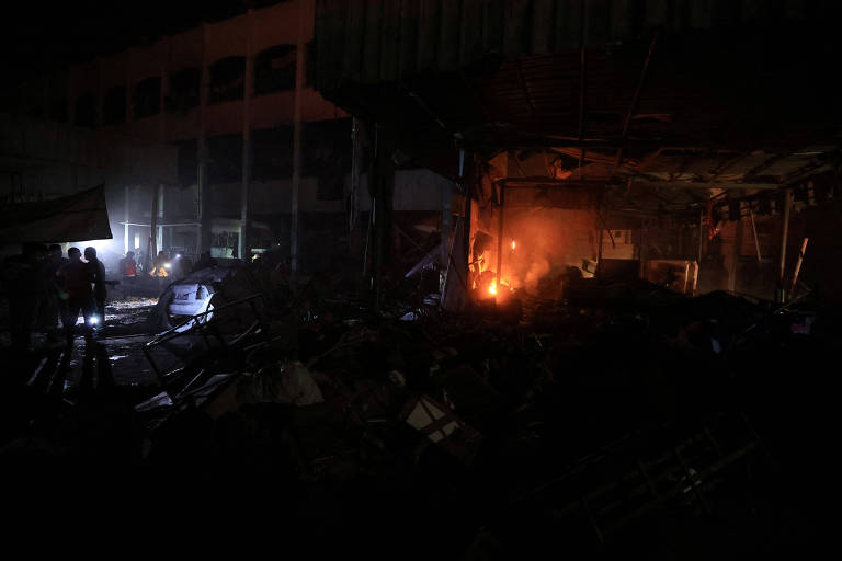 A imagem mostra um ambiente escuro e caótico, possivelmente após uma explosão ou incêndio. Há destroços espalhados pelo chão, com uma iluminação fraca proveniente de chamas e lanternas. Algumas silhuetas de pessoas podem ser vistas, aparentemente investigando a cena. O fundo é composto por estruturas danificadas e fumaça, criando uma atmosfera de desolação.