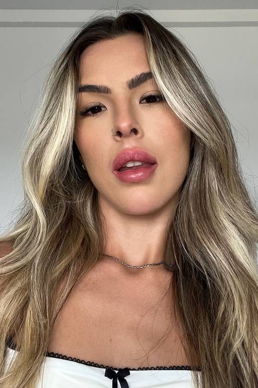 A imagem mostra uma mulher com cabelo longo e liso, em um tom loiro. Ela está usando uma blusa branca com detalhes em preto e está olhando diretamente para a câmera. O fundo é neutro e desfocado, destacando seu rosto.