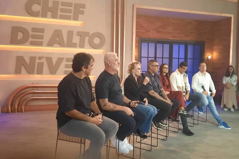 A imagem mostra um painel de seis pessoas sentadas em cadeiras em um estúdio de gravação. Ao fundo, há uma parede com o texto 'CHEF DE ALTO NÍVEL' em letras grandes. As pessoas estão vestidas de maneira casual e parecem estar participando de uma discussão ou apresentação. O ambiente é bem iluminado e moderno, com detalhes em madeira e metal.