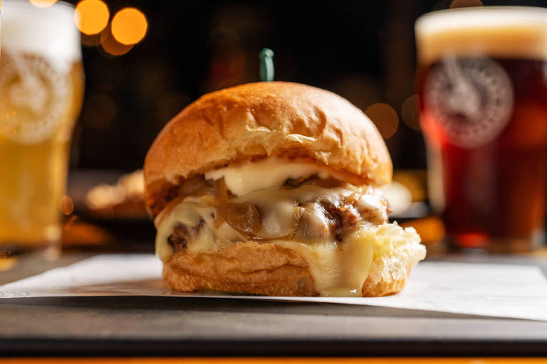 Um hambúrguer com queijo derretido e cebolas caramelizadas, servido em um pão macio. Ao fundo, há copos de cerveja, um com líquido claro e outro com líquido escuro, ambos com logotipos visíveis. A iluminação é suave, criando um ambiente acolhedor.