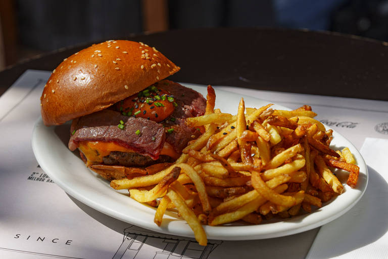 Um prato com um hambúrguer em um pão de gergelim, que contém carne, queijo e bacon, acompanhado de uma porção de batatas fritas crocantes. O prato está sobre uma mesa escura, com um fundo desfocado.