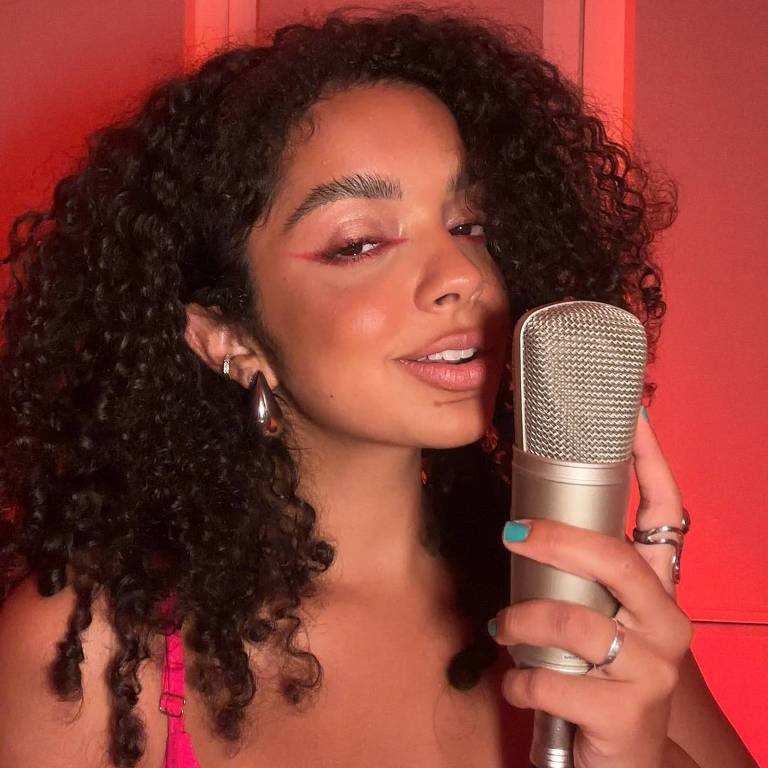 Uma jovem mulher com cabelo cacheado e volumoso, usando uma blusa rosa. Ela está segurando um microfone prateado e sorrindo para a câmera. O fundo é iluminado em tons de vermelho, criando uma atmosfera vibrante e artística.