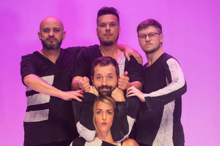 Cinco dançarinos estão em uma pose de grupo, com um fundo rosa. Eles estão vestidos com roupas pretas e brancas, formando uma composição visual interessante. Um dançarino está agachado na frente, enquanto os outros estão posicionados atrás dele, criando uma pirâmide humana. Todos têm expressões focadas e estão em uma pose dinâmica.