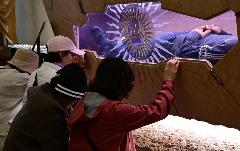 A imagem mostra três pessoas em um momento de contemplação ou oração, inclinadas em direção a um objeto ou figura visível através de um vidro. O ambiente parece ser um local sagrado ou de culto, com uma iluminação suave. Ao fundo, é possível ver uma representação simbólica com a inscrição 'IHS', que é um monograma de Jesus Cristo. As pessoas estão vestidas de maneira casual e parecem estar em um estado de reflexão.