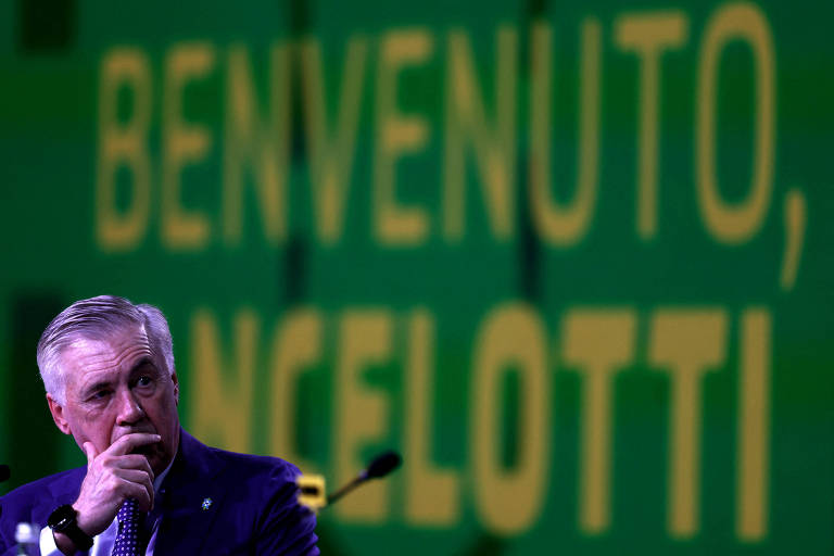 A imagem mostra um homem de cabelo grisalho sentado em uma mesa, com uma expressão pensativa. Ao fundo, há um grande painel com a inscrição em letras grandes e amarelas: 'BENVENUTO, ANCELOTTI'. O ambiente parece ser uma conferência ou apresentação, com microfones na mesa.