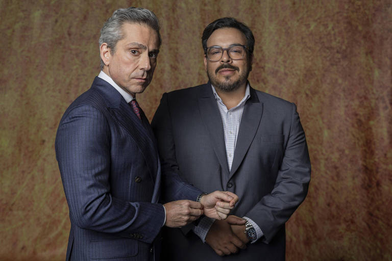 A imagem mostra dois homens em pé, um ao lado do outro. O homem à esquerda está usando um terno escuro com listras e uma gravata vermelha, enquanto o homem à direita está vestido com um terno cinza e uma camisa clara. Ambos têm expressões sérias e estão posando para a foto. O fundo da imagem é de uma cor marrom avermelhada.