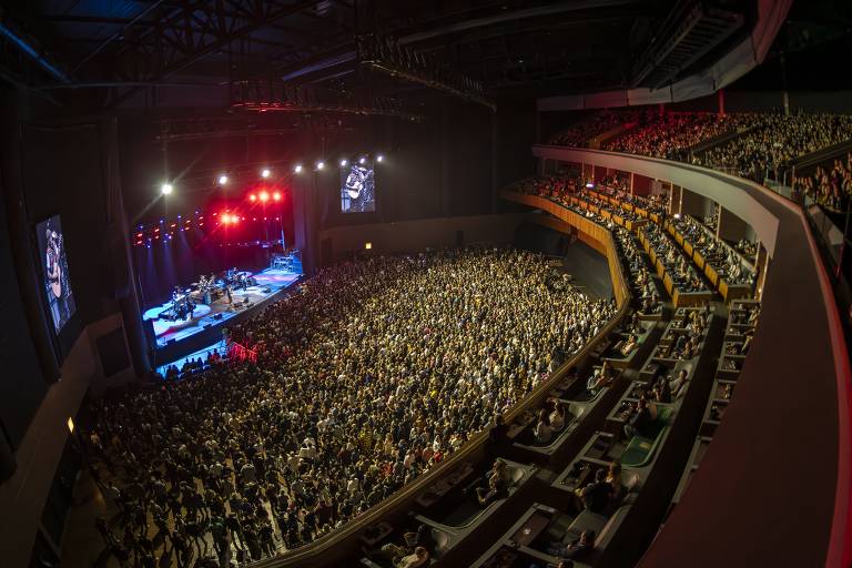 A imagem mostra um grande auditório lotado durante um show. O palco está iluminado com luzes vermelhas e brancas, e há uma banda tocando. O público é visível em várias seções, com muitas pessoas em pé e aplaudindo. As arquibancadas estão cheias, e há telões visíveis ao lado do palco.