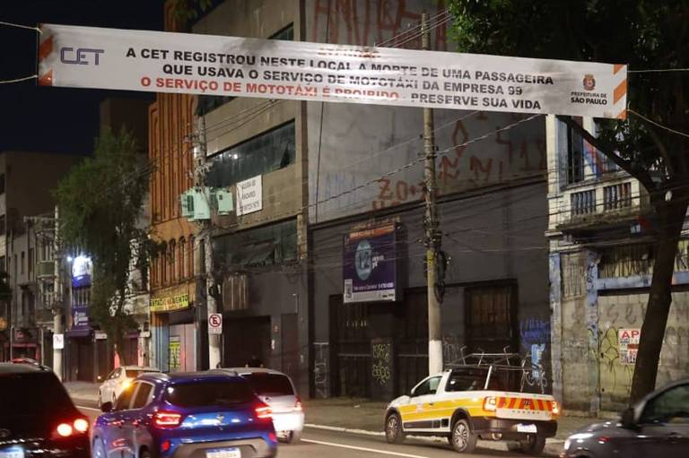 A imagem mostra uma rua à noite com vários carros em movimento. À esquerda, há um carro preto e um carro azul, enquanto à direita, um táxi amarelo está estacionado. No fundo, há um banner suspenso que diz: 'A CET registrou neste local a morte de uma passageira que usava o serviço de mototáxi da empresa 99. O serviço de mototáxi é perigoso, preserva sua vida.' O ambiente é urbano, com prédios e iluminação noturna visíveis.