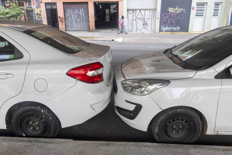 A imagem mostra dois carros brancos estacionados muito próximos um do outro, com os para-choques traseiro e dianteiro quase se tocando. O carro da esquerda tem um pneu sobressalente visível, enquanto o carro da direita possui um design mais moderno. Ao fundo, há uma rua com grafites nas paredes e uma pessoa caminhando.