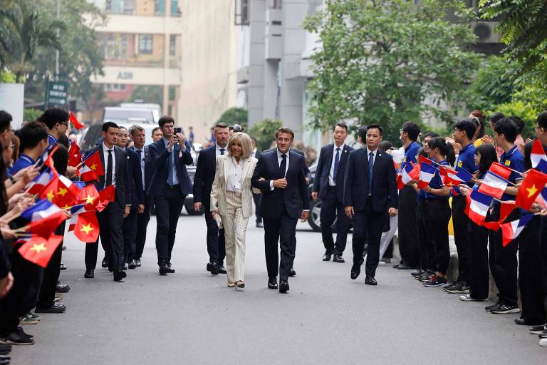 A imagem mostra uma recepção oficial em uma rua, onde um grupo de pessoas está segurando bandeiras do Vietnã e de outro país. No centro, um homem e uma mulher caminham juntos, acompanhados por um grupo de seguranças e assessores. O ambiente é urbano, com prédios ao fundo e árvores ao lado da rua.