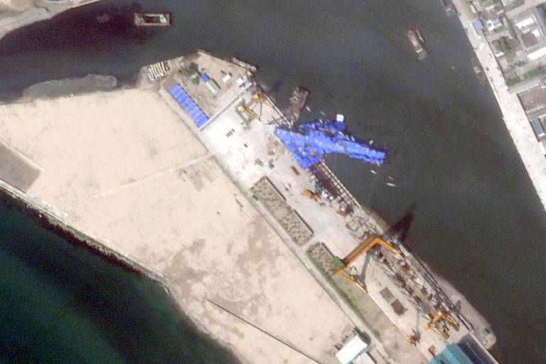 A imagem mostra uma vista aérea de um porto. É possível ver uma área de cais com estruturas de metal e equipamentos de carga. Há também barcos no canal próximo ao cais. A água é de cor escura e há uma faixa de areia clara na parte inferior da imagem. O céu parece limpo e a área ao redor do porto é urbanizada, com edifícios visíveis ao fundo.