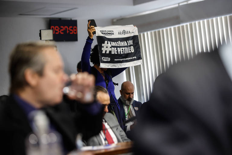 A imagem mostra uma reunião onde uma pessoa está segurando um cartaz com a hashtag "#MarianaLimeDePetroleo". Ao fundo, há um relógio digital que marca o tempo e várias pessoas sentadas, algumas delas visíveis em primeiro plano. A iluminação é natural, proveniente de janelas com persianas.