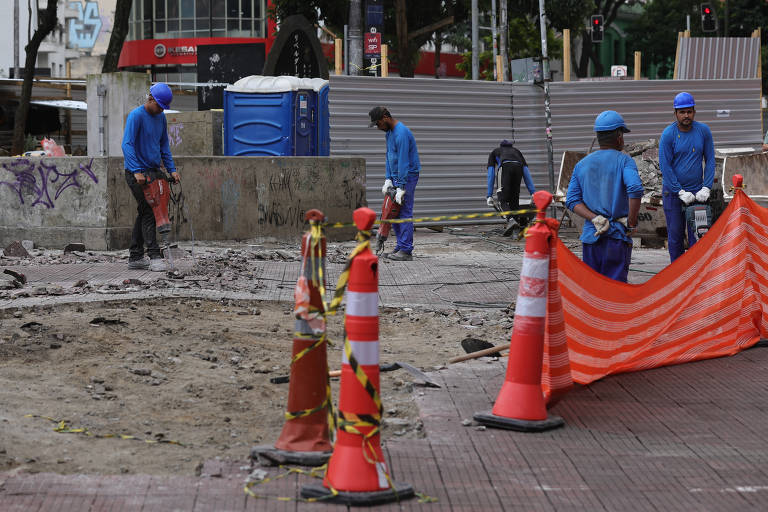 A imagem mostra um grupo de trabalhadores em uma área de construção. Eles estão vestidos com uniformes azuis e capacetes, realizando atividades em um local onde o pavimento foi removido. Há cones de sinalização laranja e uma barreira de segurança laranja ao fundo, indicando que a área está em obras. No fundo, é possível ver uma cabine de banheiro químico e estruturas urbanas.