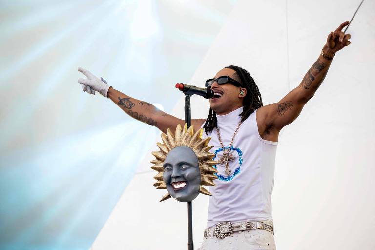 Um artista se apresenta em um palco, usando uma camiseta branca e calças brancas. Ele tem cabelo longo e usa óculos escuros. O artista está segurando um microfone e gesticulando com a outra mão. Ao lado dele, há um suporte com uma máscara de sol sorridente. O fundo é claro, com luzes que criam um efeito dramático.