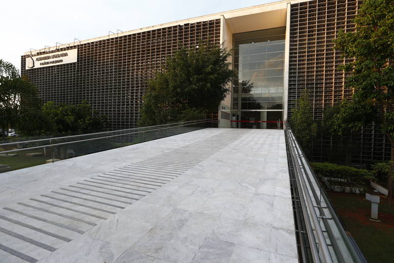 A imagem mostra a fachada da Assembleia Legislativa, com uma rampa de acesso em mármore e um grande painel de vidro na entrada. O edifício possui uma estrutura moderna, com uma grade de madeira ou metal em sua parte superior. Ao redor, há áreas verdes com árvores e arbustos.