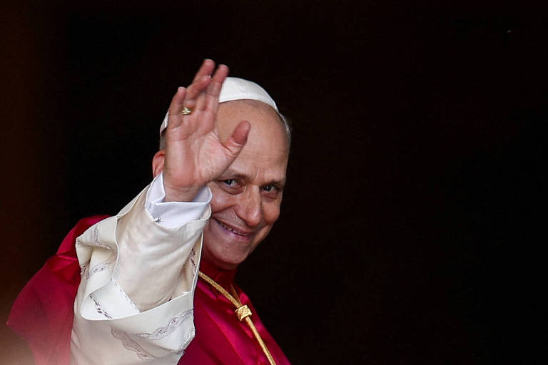 Um homem vestido com vestes papais, incluindo uma túnica vermelha e uma capa branca, está acenando com a mão direita. Ele tem um sorriso no rosto e está em um ambiente escuro, possivelmente uma sacada ou janela. O fundo é indistinto, mas sugere um cenário solene.