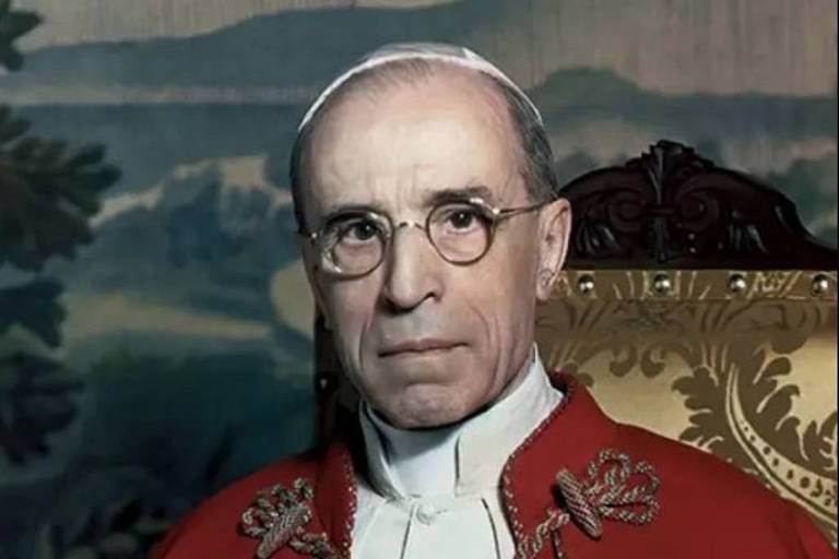 A imagem mostra um clérigo idoso vestindo uma batina branca com uma capa vermelha e um colar com um crucifixo. Ele está sentado com as mãos cruzadas e usa óculos. O fundo da imagem é desfocado, mas parece ser uma paisagem com árvores e montanhas.