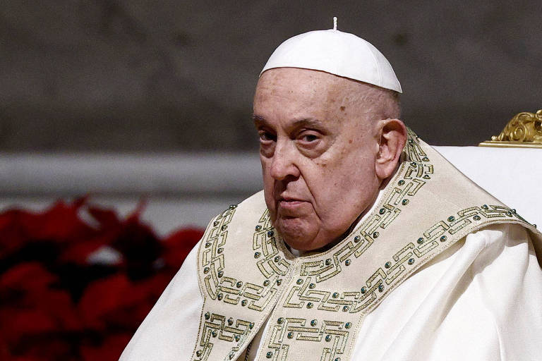Um homem idoso com cabelo branco e pele clara, usando uma vestimenta religiosa branca e dourada, está sentado em uma cadeira. Ele tem uma expressão séria e está olhando para frente. Ao fundo, há uma decoração de Natal com folhas vermelhas.