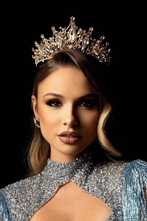 A imagem mostra uma mulher com uma coroa elaborada na cabeça. Ela está vestindo um traje brilhante com detalhes em pedrarias e franjas. O fundo é preto, destacando seu rosto e a coroa. A mulher tem cabelo loiro ondulado e usa maquiagem que realça seus olhos e lábios.