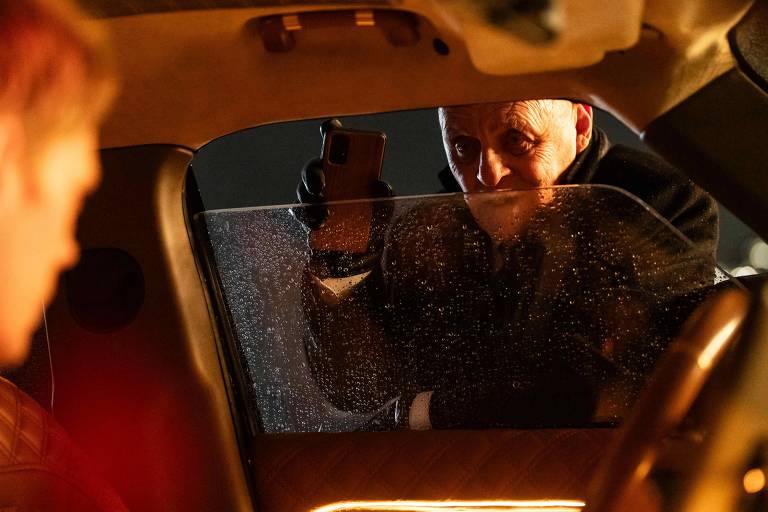 A imagem mostra um homem dentro de um carro, visivelmente nervoso, enquanto um outro homem, do lado de fora, se aproxima com um celular na mão. A janela do carro está embaçada, e a cena parece ocorrer à noite, com iluminação suave. O homem do lado de fora está usando luvas e tem uma expressão séria, sugerindo uma situação tensa.