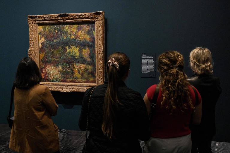 A imagem mostra um grupo de quatro pessoas observando uma pintura emoldurada, que está pendurada em uma parede de cor escura. As pessoas estão de costas, voltadas para a obra de arte, e parecem estar apreciando a pintura. A moldura da pintura é elaborada e dourada, e ao lado dela há uma placa informativa sobre a obra.